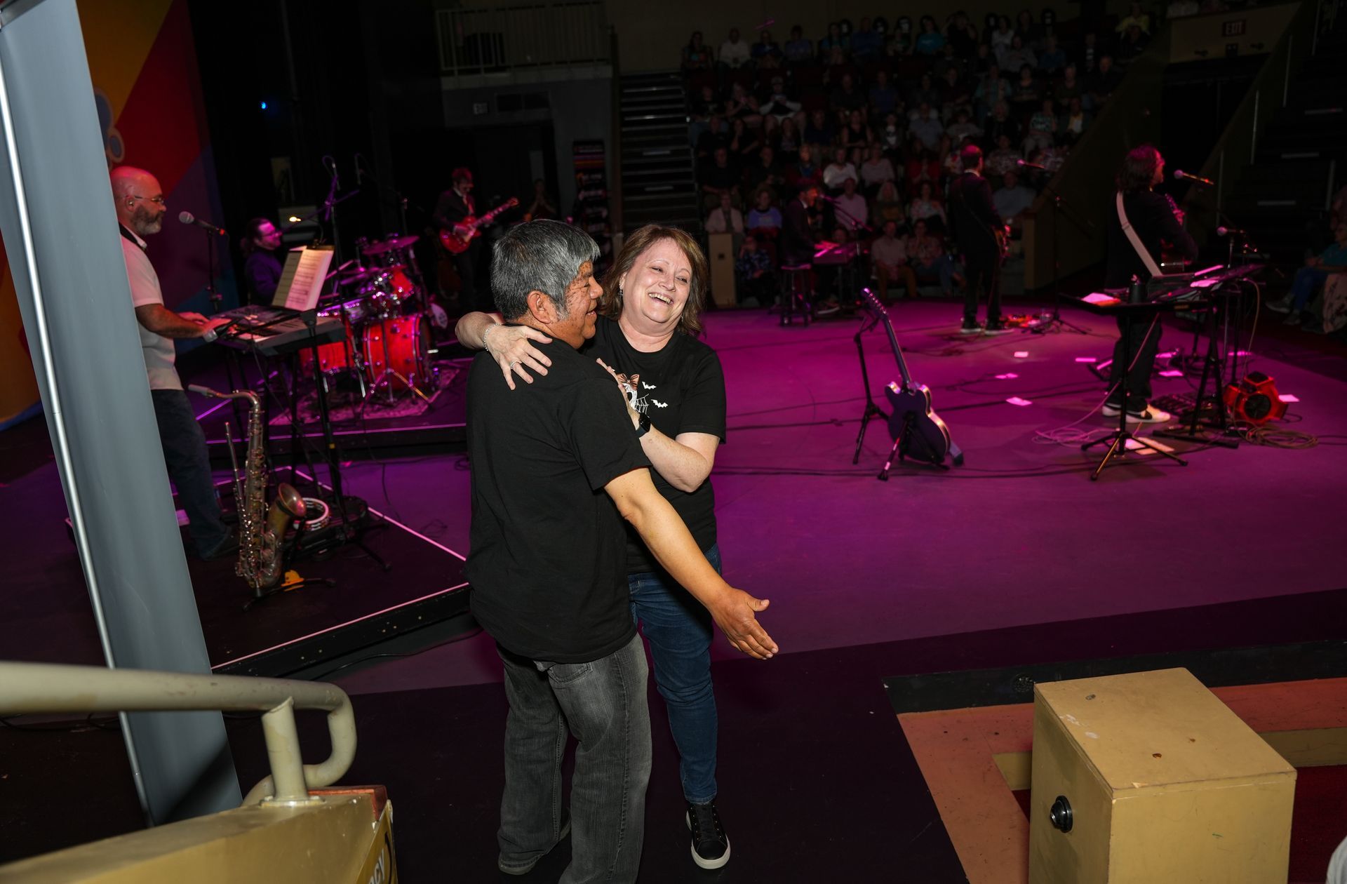 A man and woman embrace onstage at a concert, smiling. The band performs in the background.
