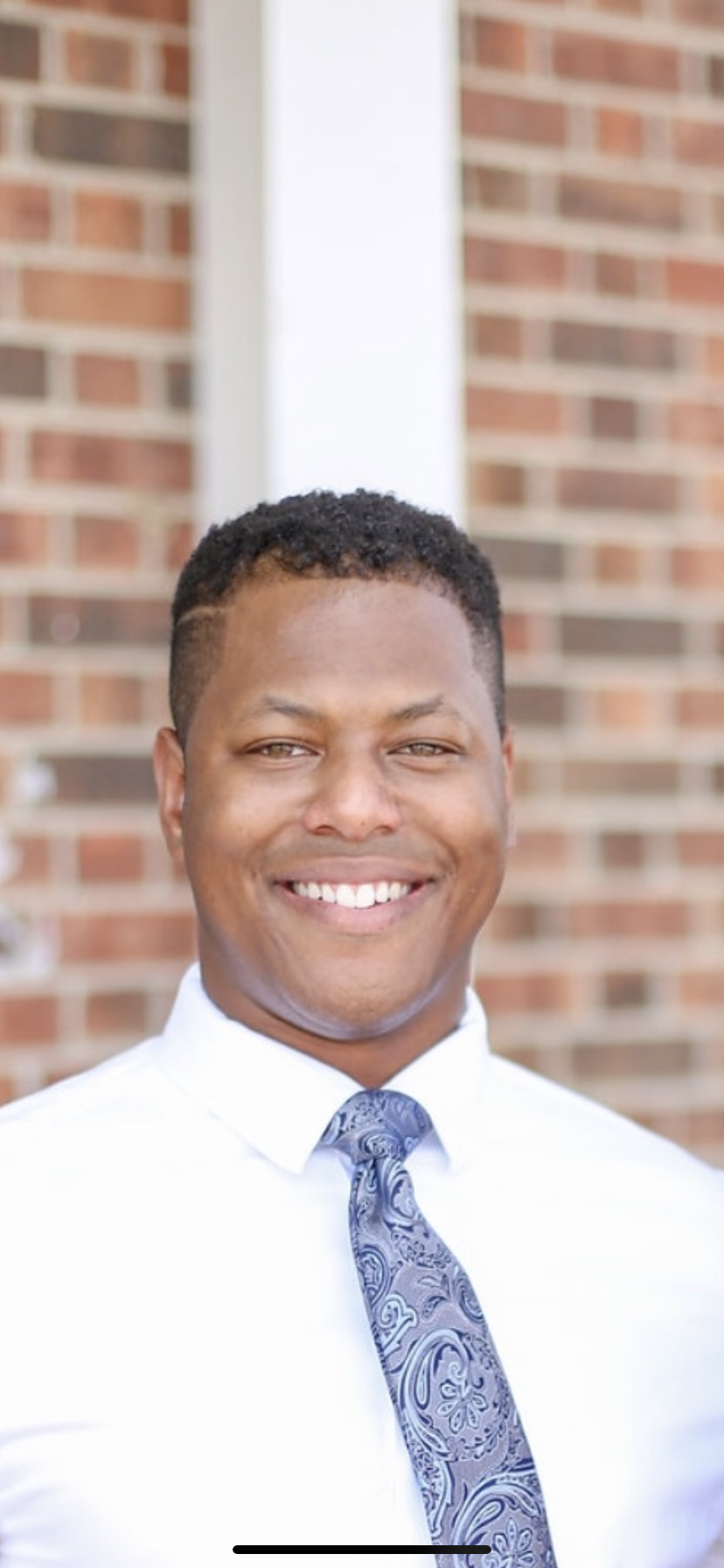 A smiling Black man in a white shirt and patterned tie, posed in front of a brick wall.
