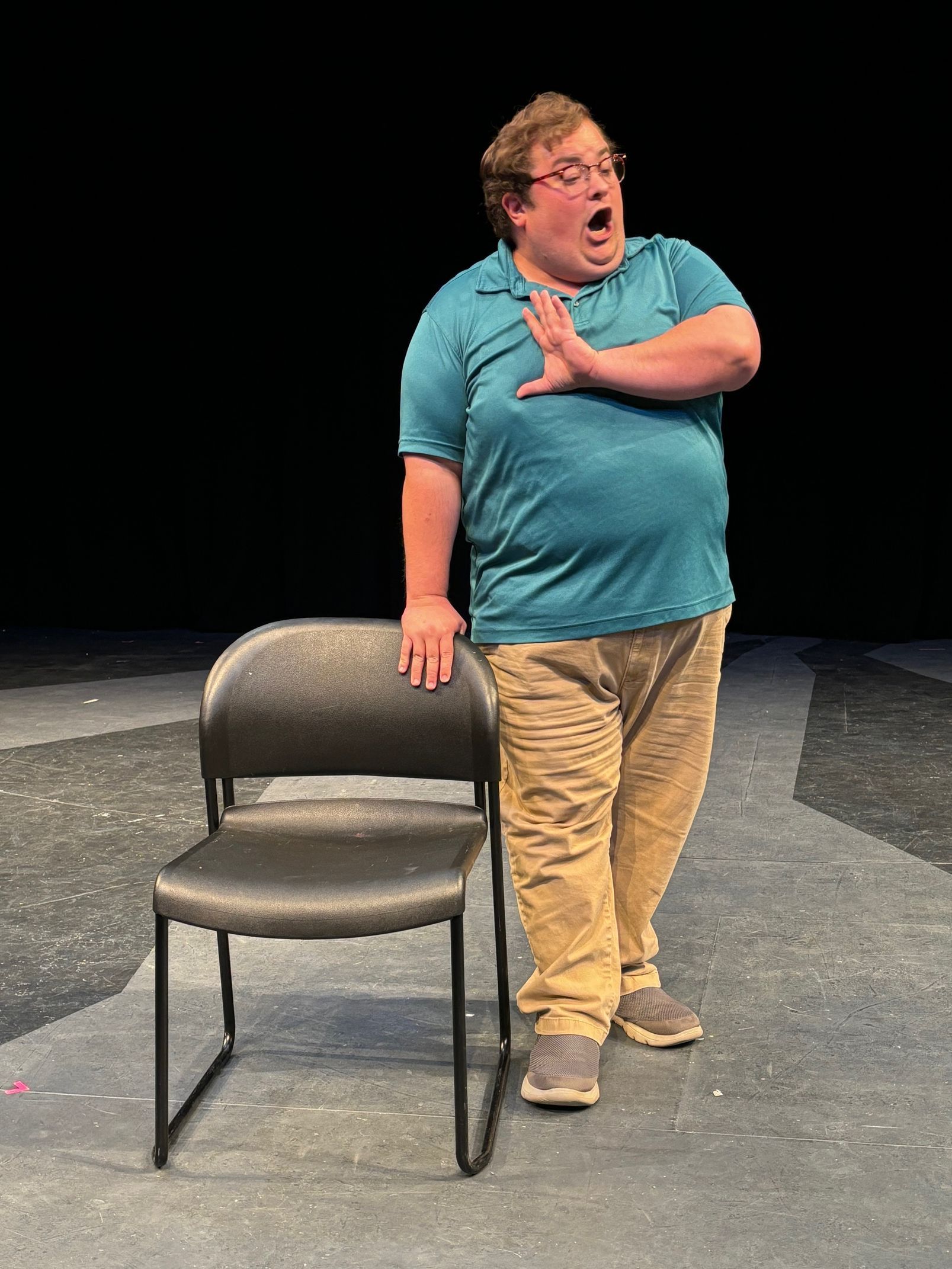 Man in teal shirt and khakis singing on stage, leaning on a black chair, hand raised.