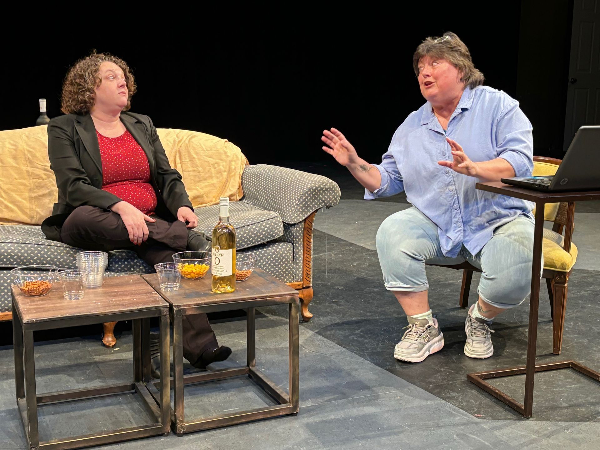 Two women in a play: One on a couch, the other gesturing, sitting next to a laptop.