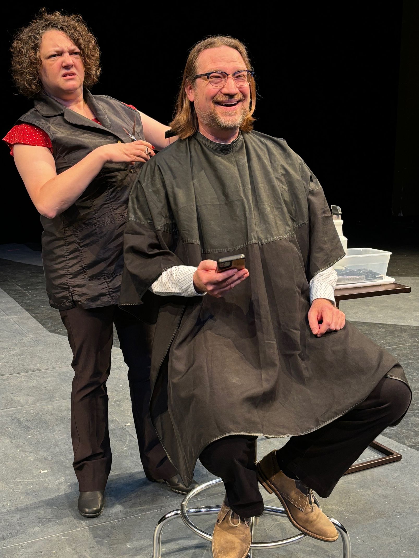 Woman giving a man a haircut. She wears a gray vest. Man smiles, looking at his phone, wearing a black cape.
