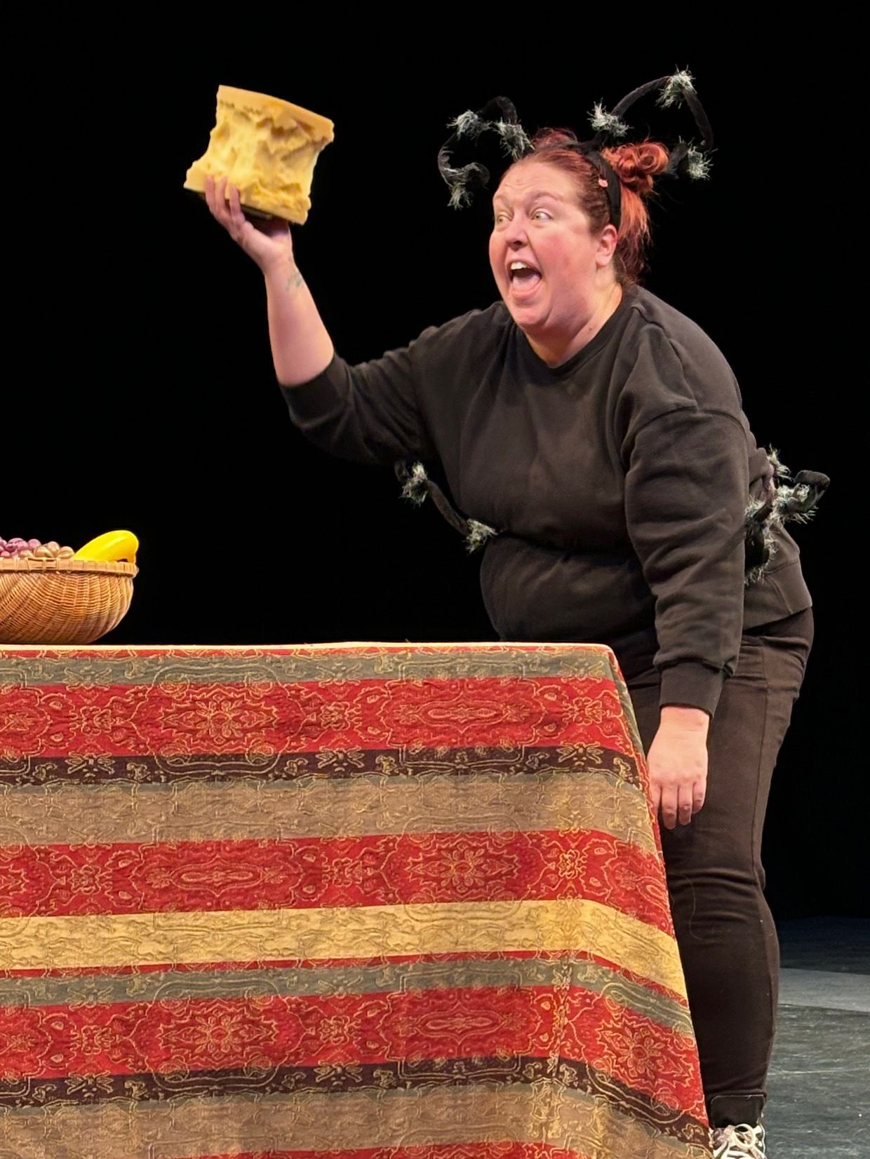 Woman with antenna headgear holds up a large bread piece, open-mouthed, beside a table with food.