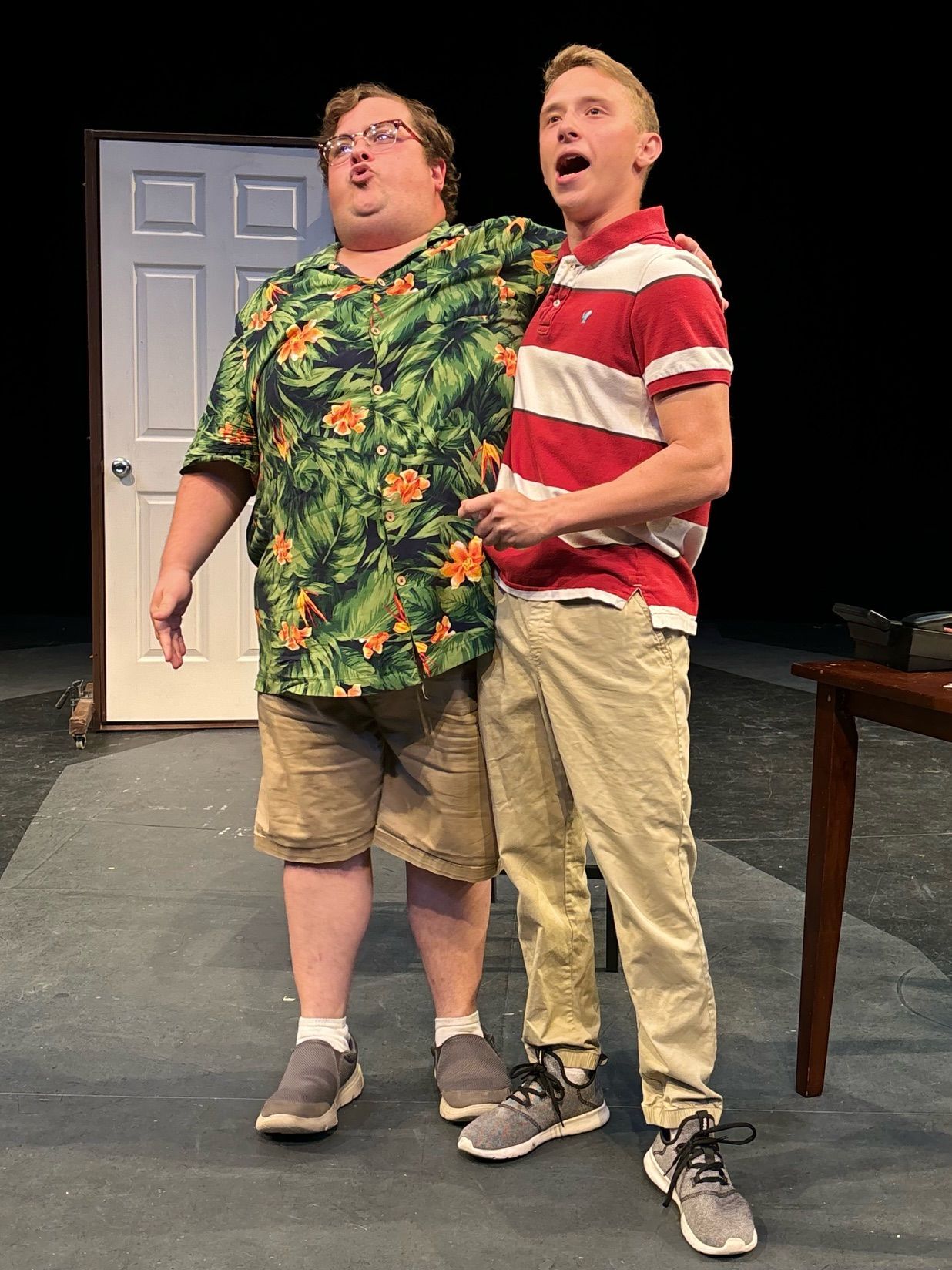 Two men on stage singing: One in a floral shirt and shorts, the other in a red striped shirt and khakis.
