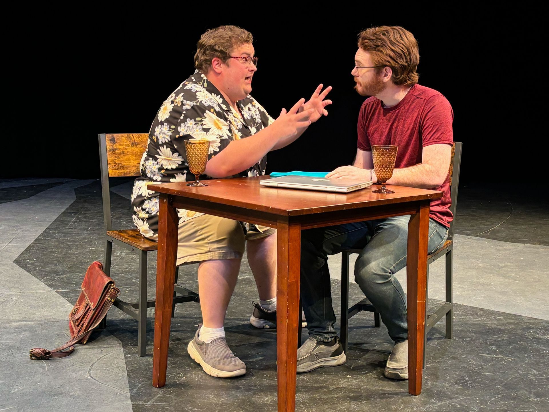 Two men seated at a table, one gesturing. Stage set: dark backdrop, table, chairs.