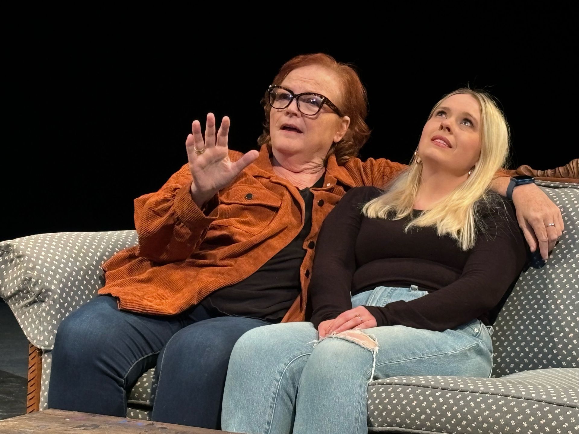 Two women on a couch: older woman gestures, young woman looks up, both are indoors.