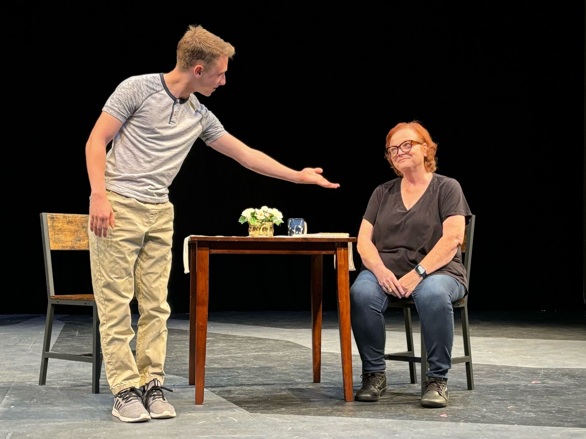 A young man gestures toward a woman seated at a table on a stage.