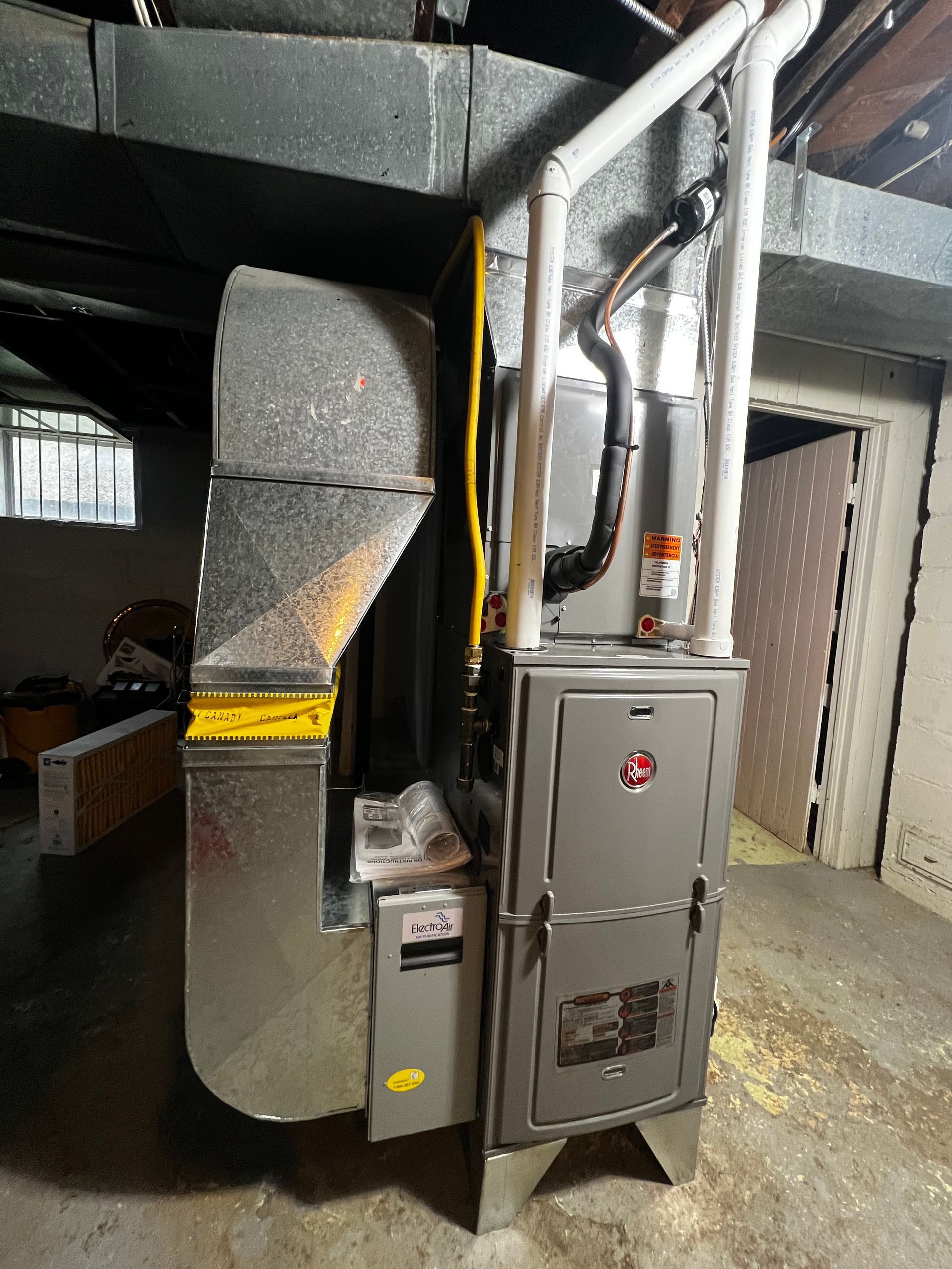 A furnace is sitting in a basement next to a window.