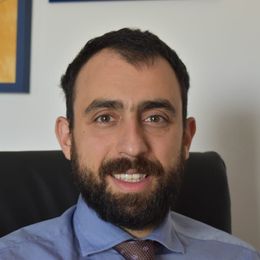 Uomo sorridente con la barba, che indossa una camicia blu e una cravatta, seduto su una sedia da ufficio.