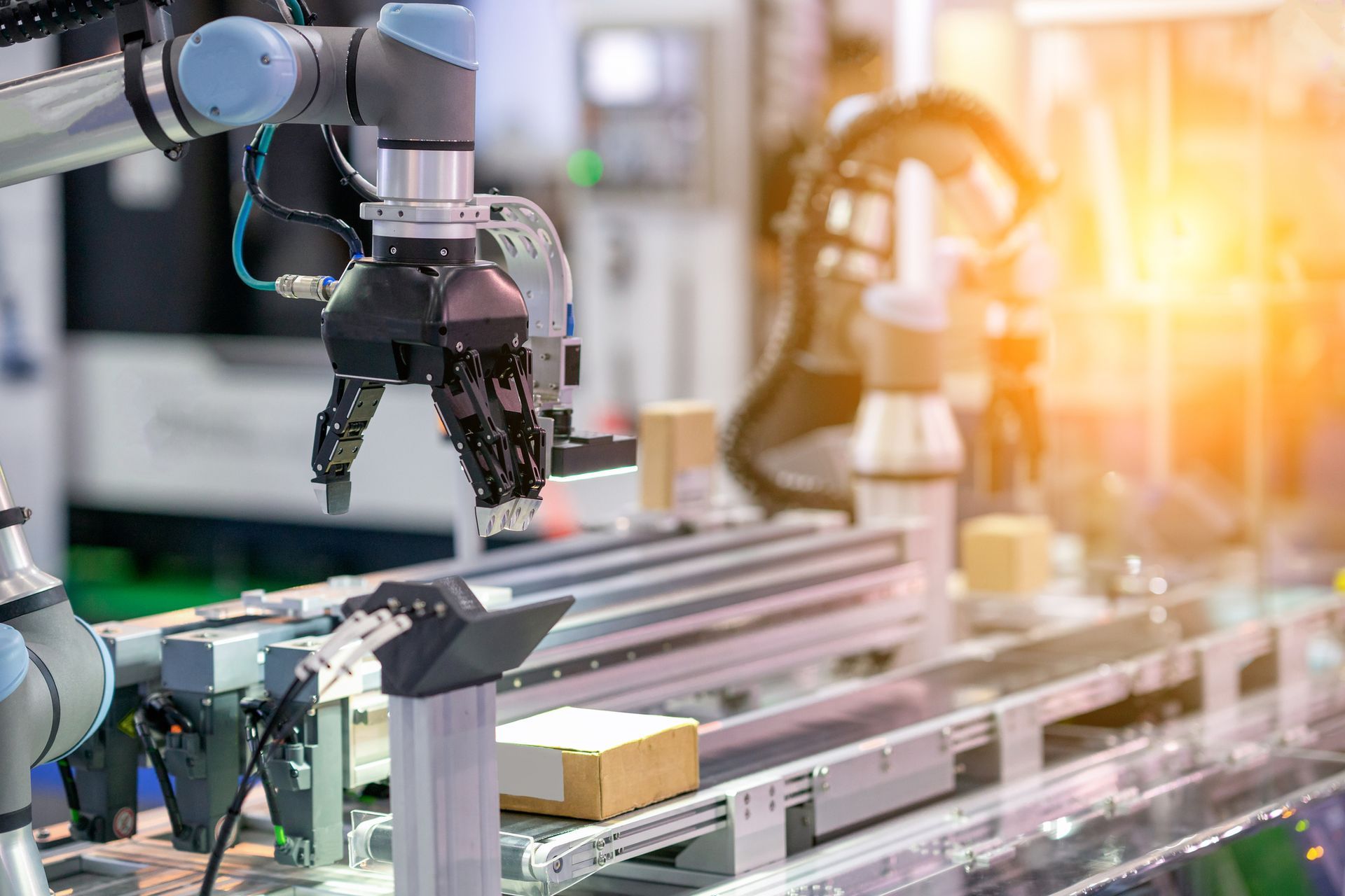 Bracci robotici su una catena di montaggio in una fabbrica, uno dei quali afferra una scatola di cartone. La luce del sole illumina la scena.