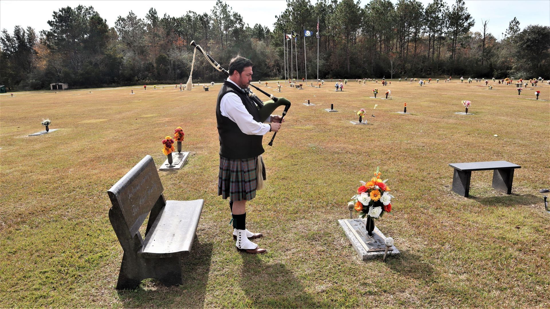 A bagpiper in a kilt plays by a headstone with flowers in a grassy cemetery.
