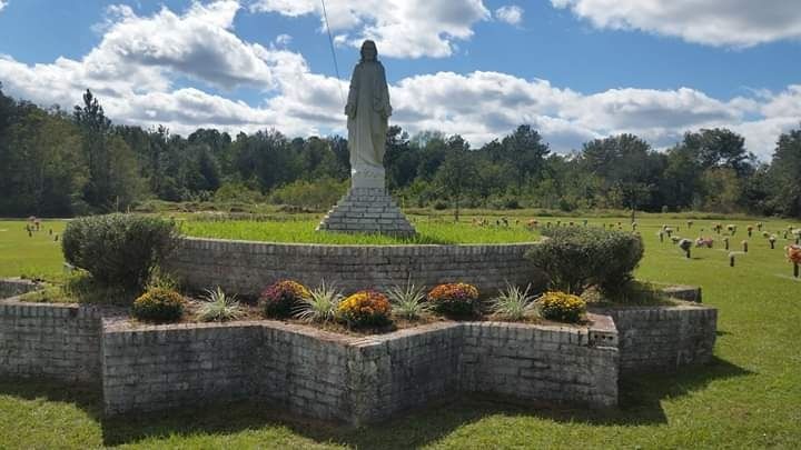 A stone statue stands atop a star-shaped brick planter in a sunny, green cemetery.