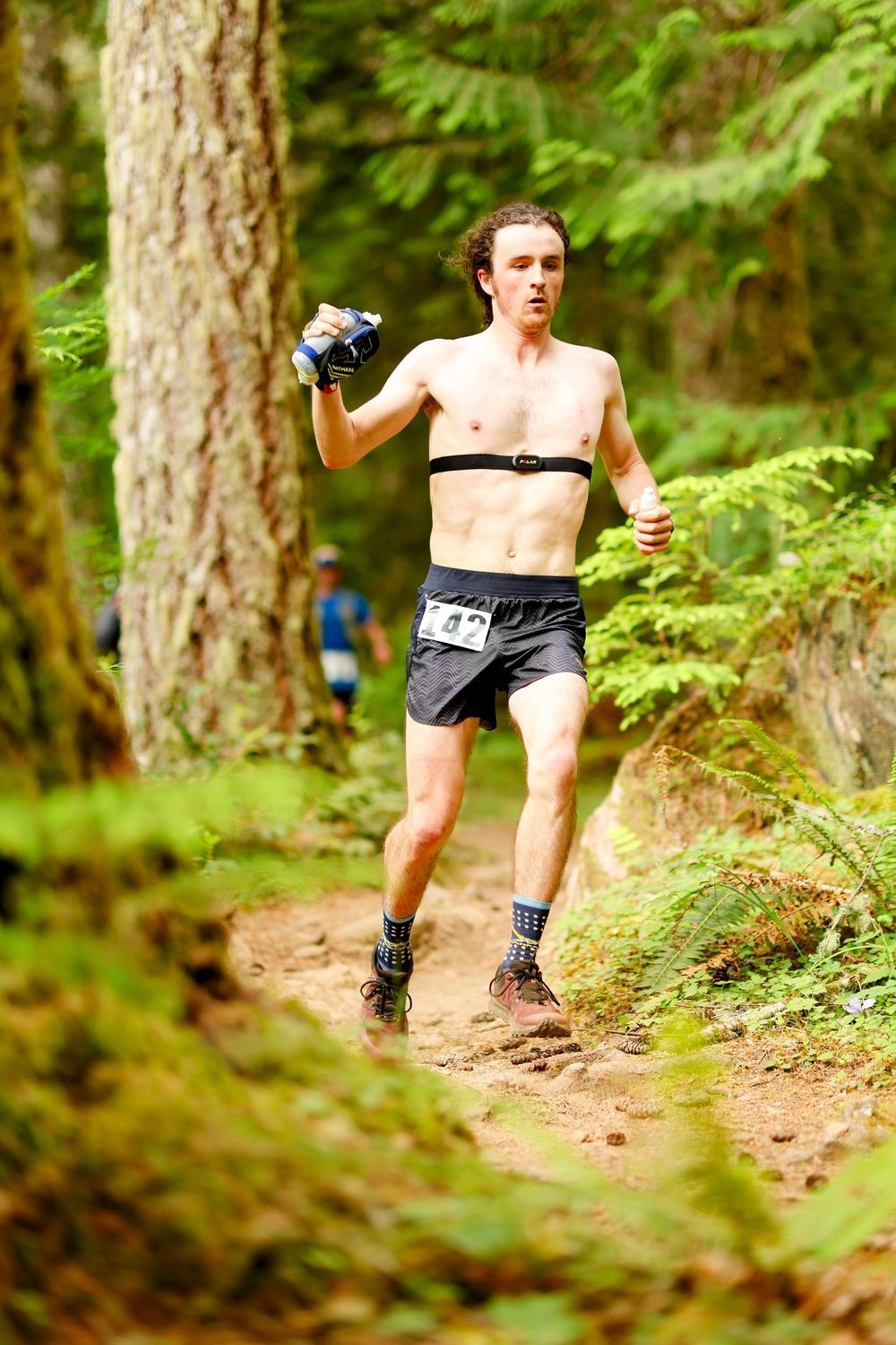 A shirtless man is running on a trail in the woods.
