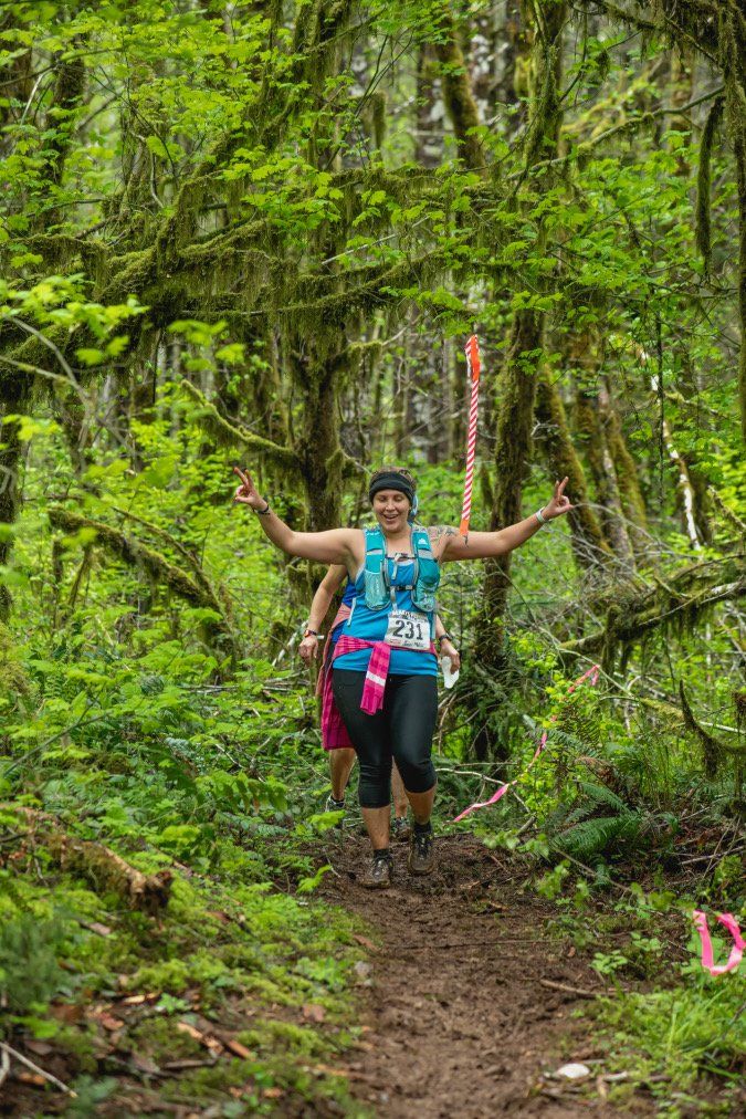 A woman is walking down a trail in the woods with her arms outstretched.