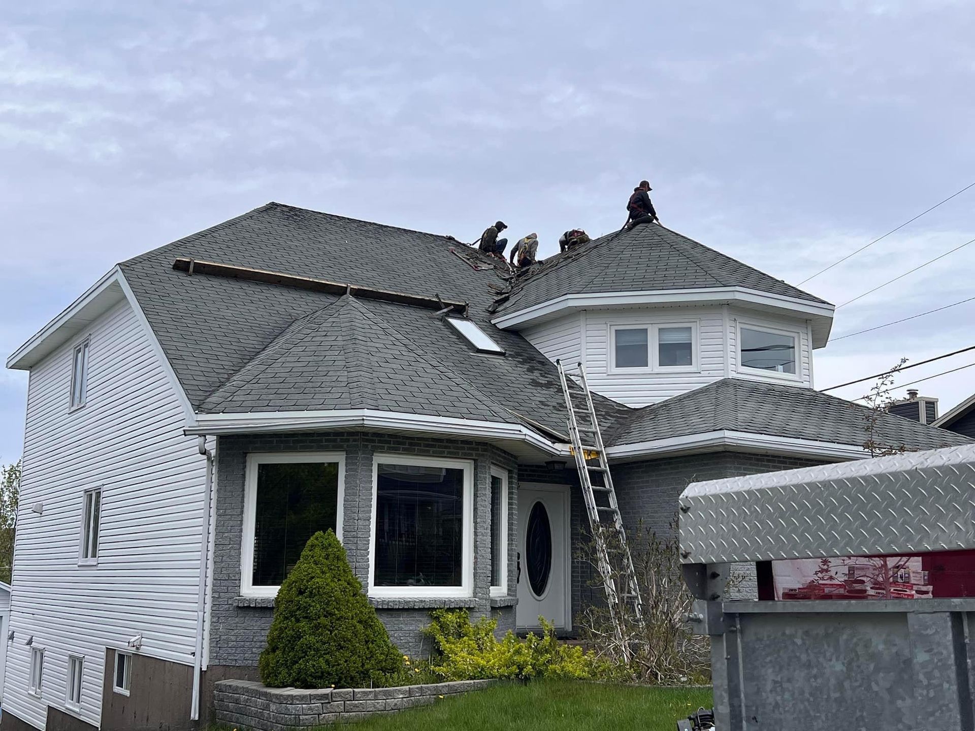 People working on a gray shingled roof of a two-story house on a cloudy day. A ladder leans against the house.