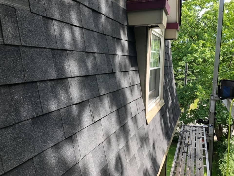 Dark gray asphalt shingles cover a steeply pitched roof with a dormer window. A scaffold is visible below.