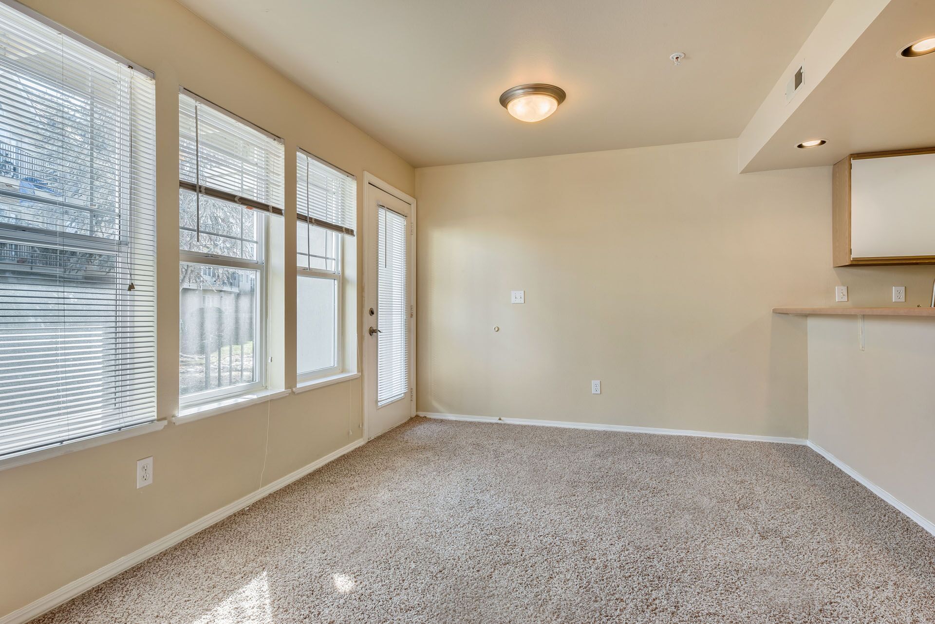 Empty beige living room with carpet, large windows, and open kitchen counter area