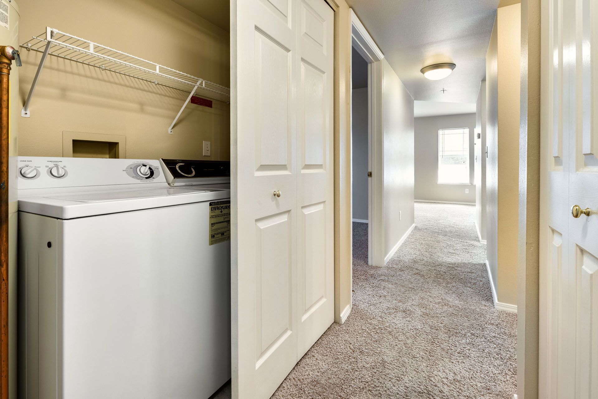 Laundry closet beside a carpeted hallway with white doors and a bright window at the end.