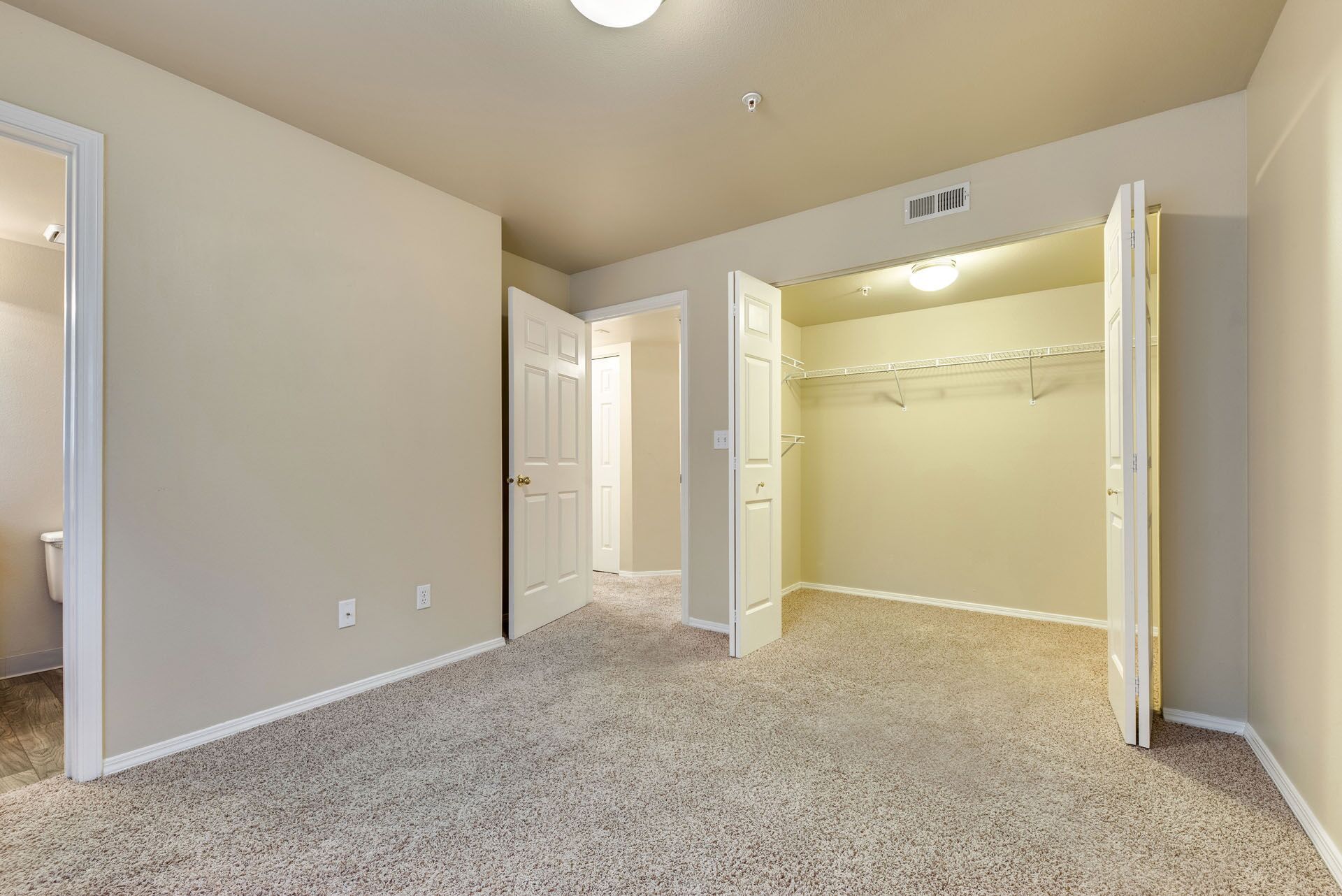 Empty beige bedroom with carpet and open closet doors, seen from the doorway.