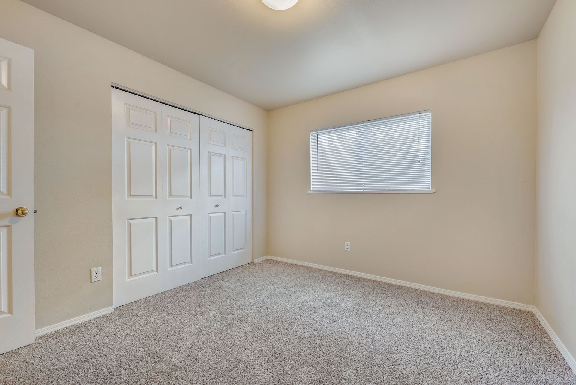 Empty beige bedroom with carpet, closet doors, and a small window with blinds.