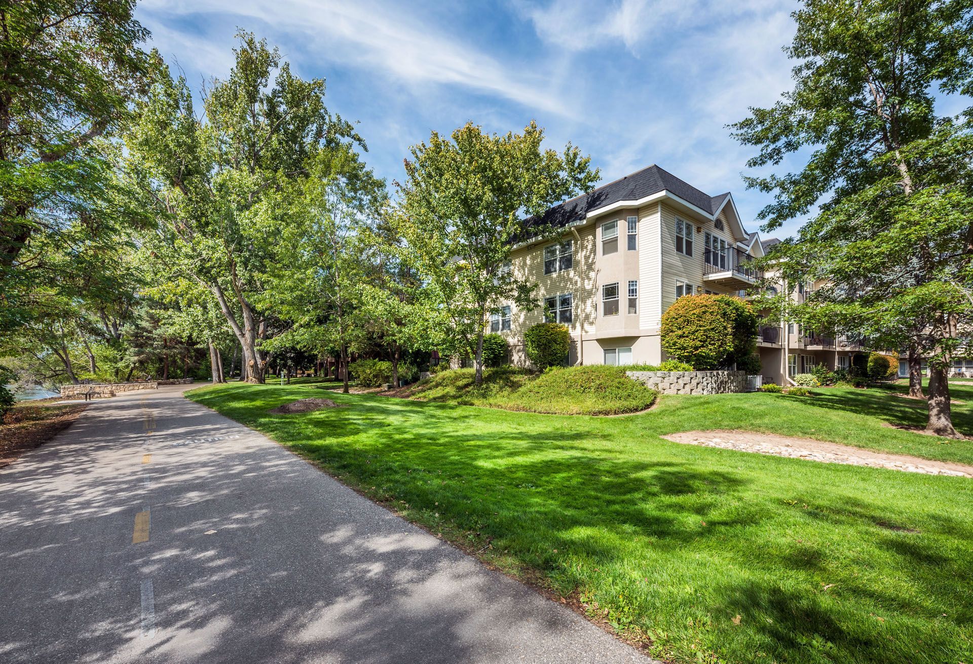 Tree-lined driveway beside a beige apartment building with green lawn and blue sky