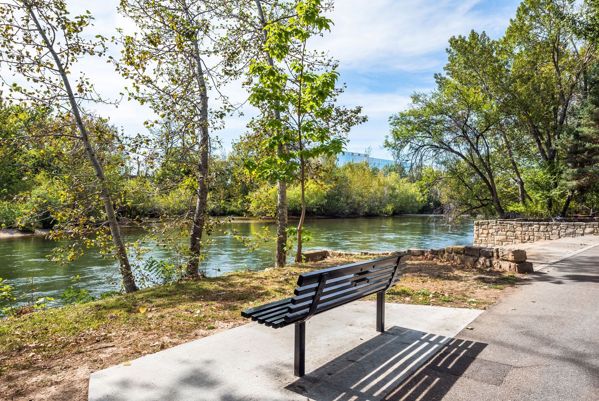 Park bench beside a sunlit river with trees and a stone path