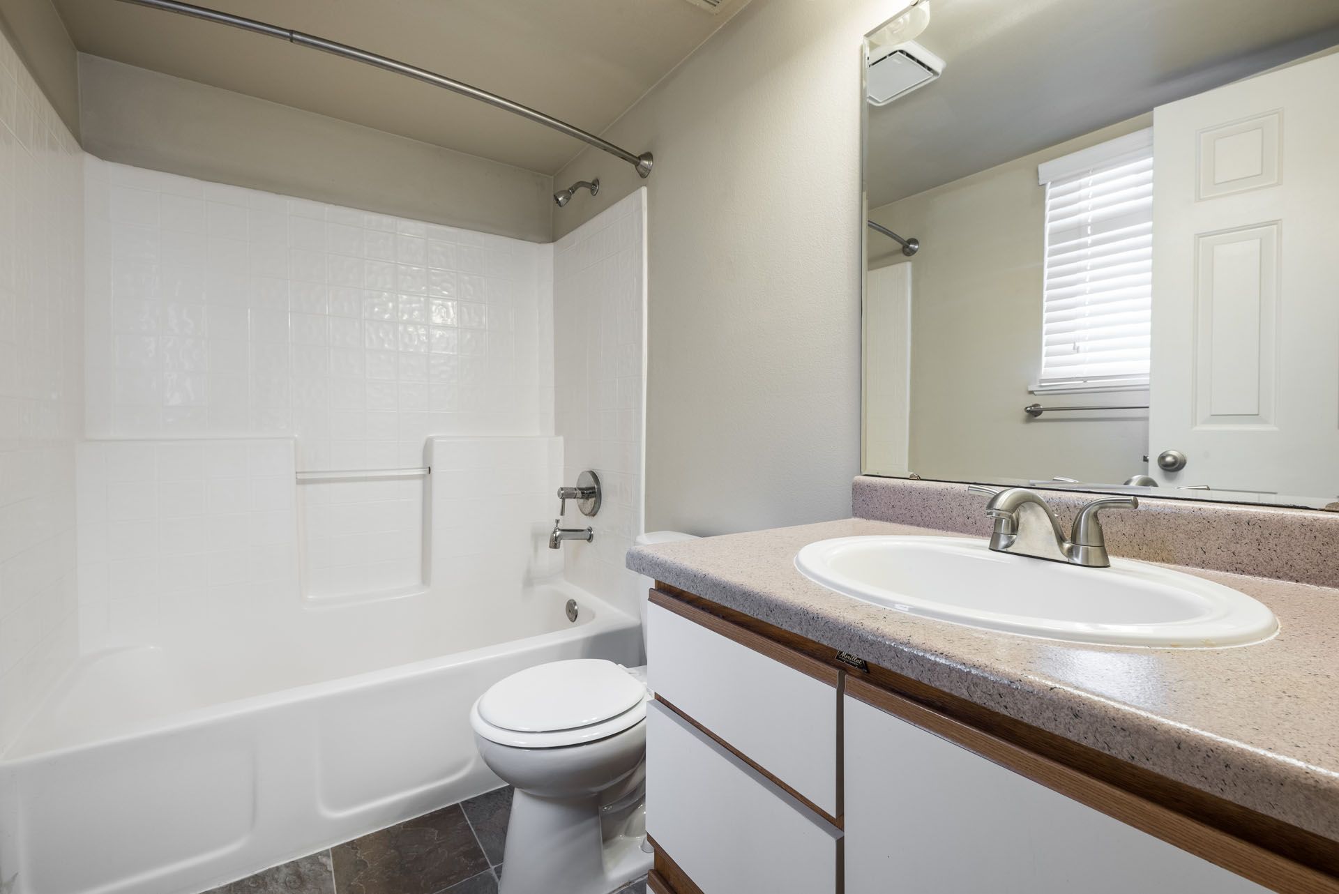 Bright bathroom with white tub and toilet, beige vanity counter, and window blinds.