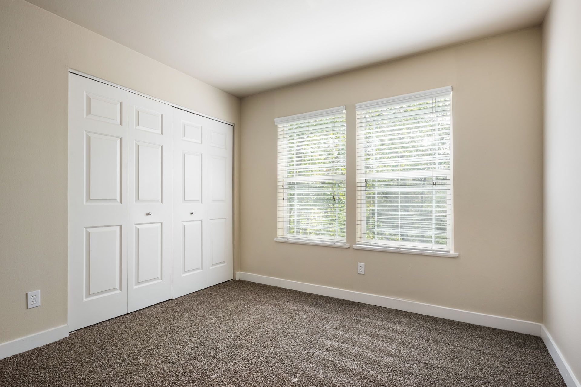 Empty beige bedroom with carpet, white closet doors, and two windows with blinds.