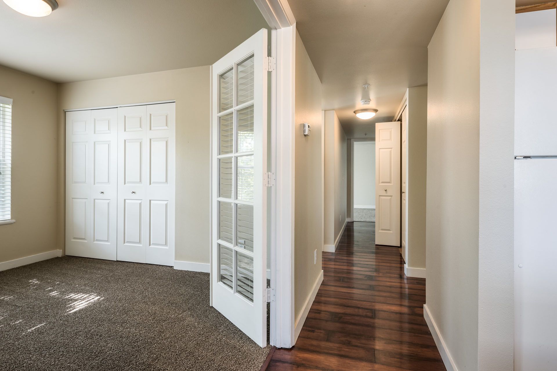 Empty beige bedroom with carpet, French door, closet doors, and a hallway with wood floors