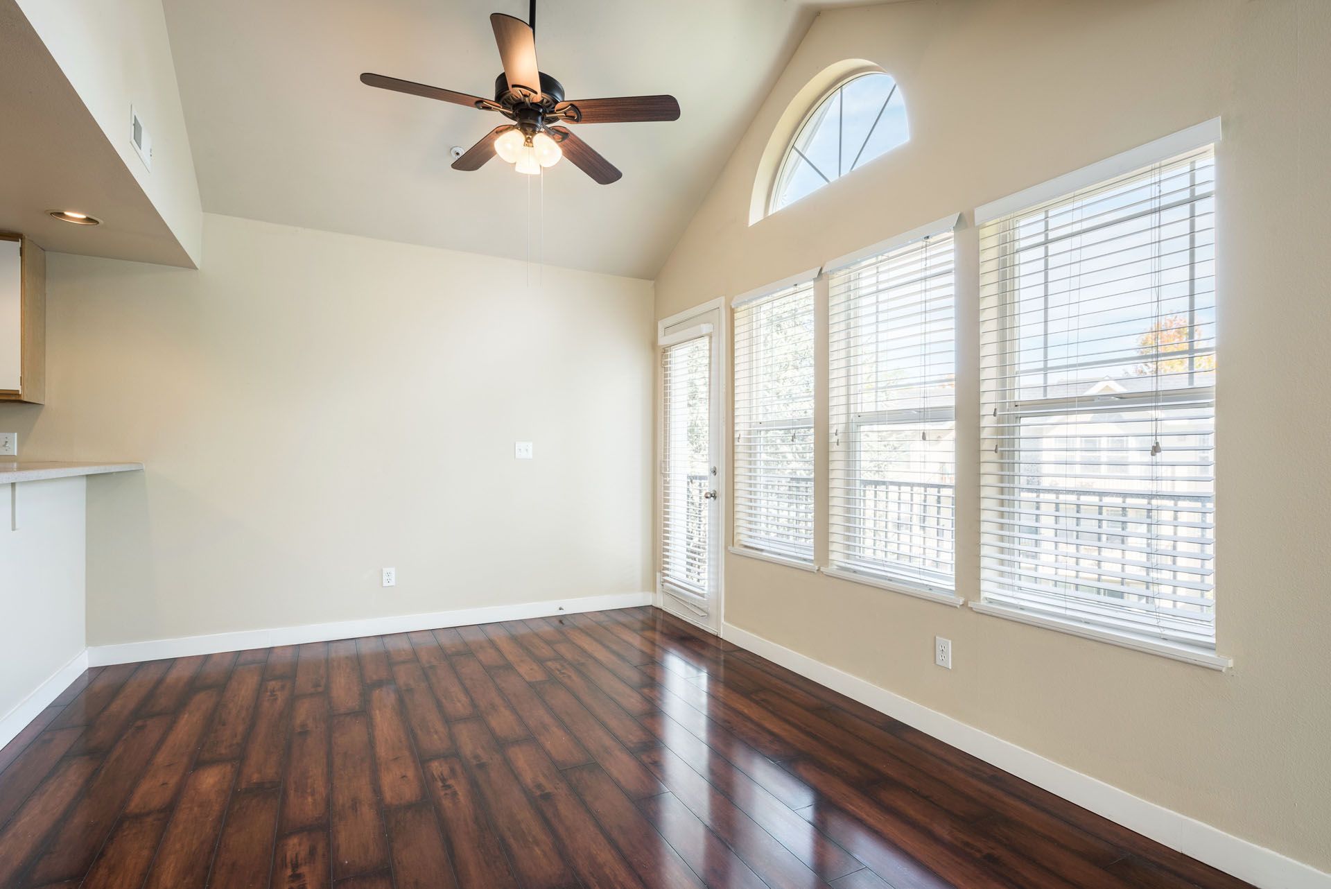 Empty living room with dark wood floors, white walls, ceiling fan, and large windows with blinds