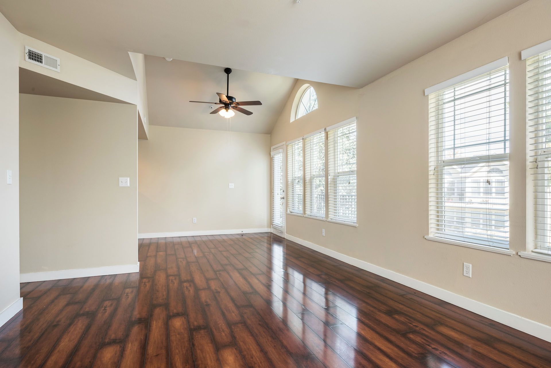Empty living room with dark wood floors, white walls, large windows, and a ceiling fan