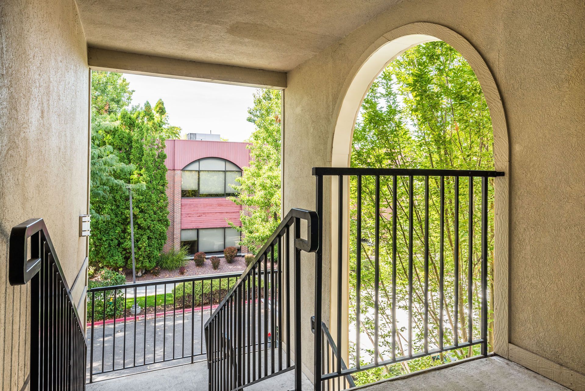Arched balcony view of a courtyard and pink building framed by green trees and black railings