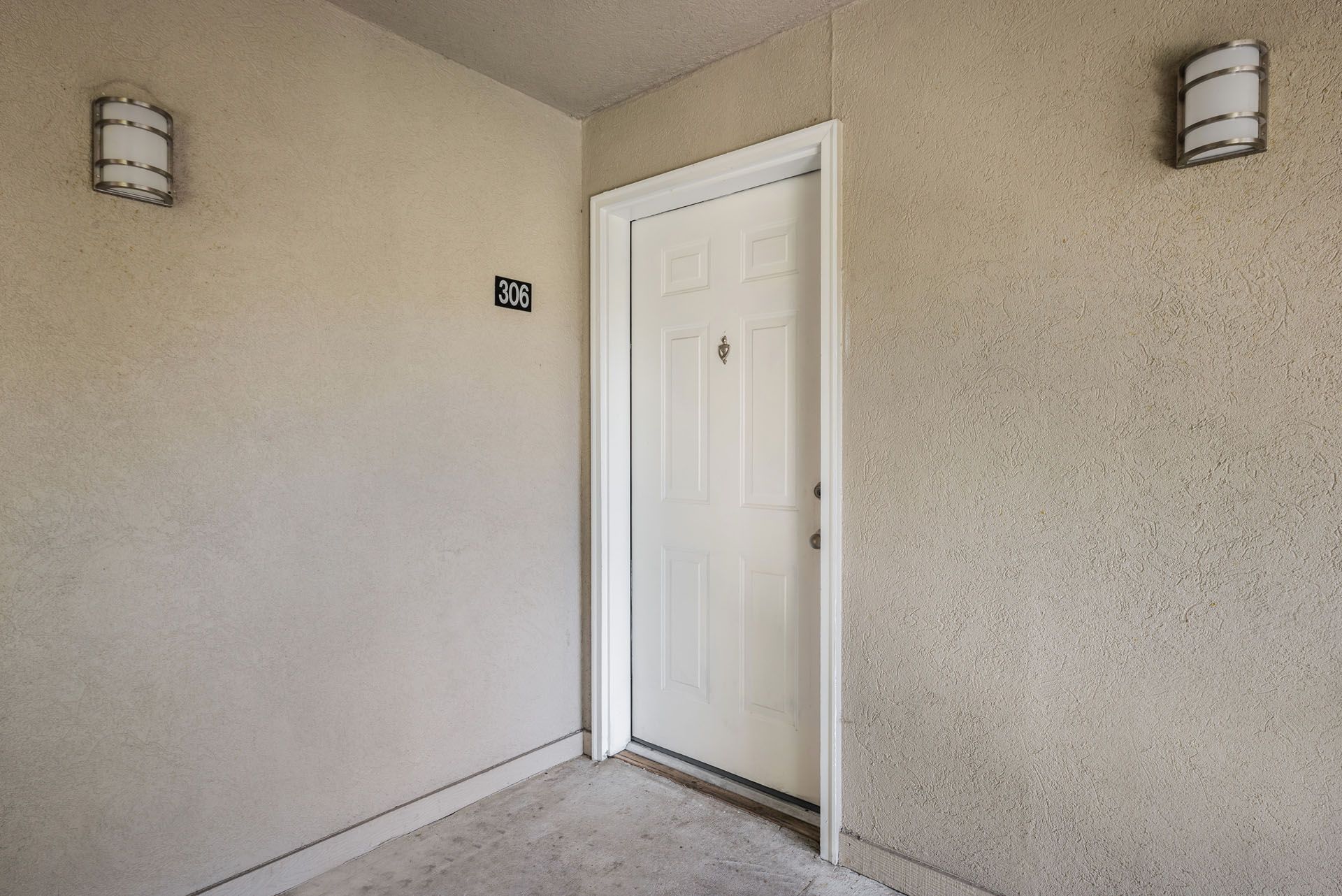 Exterior entryway with a white door and beige stucco walls, lit by two wall-mounted lights.