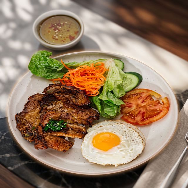 Plate of grilled meat with fried egg, shredded carrot, lettuce, cucumber, tomato, and dipping sauce on a table