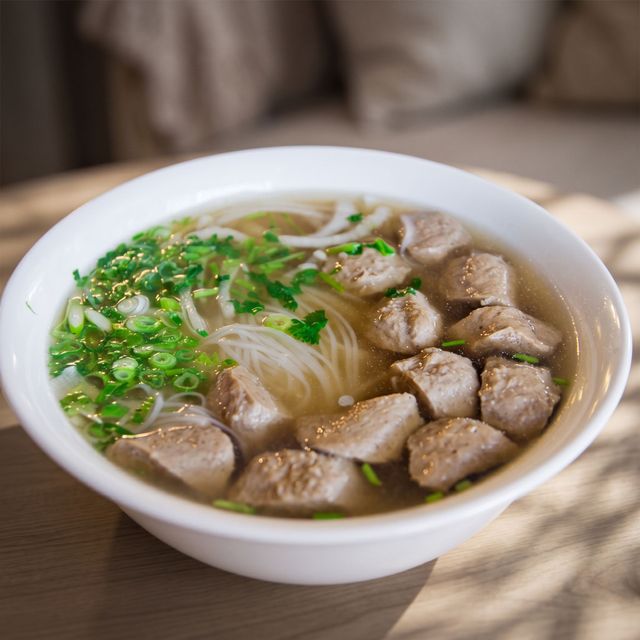 A bowl of noodle soup with sliced meat, green onions, and meatballs on a table.