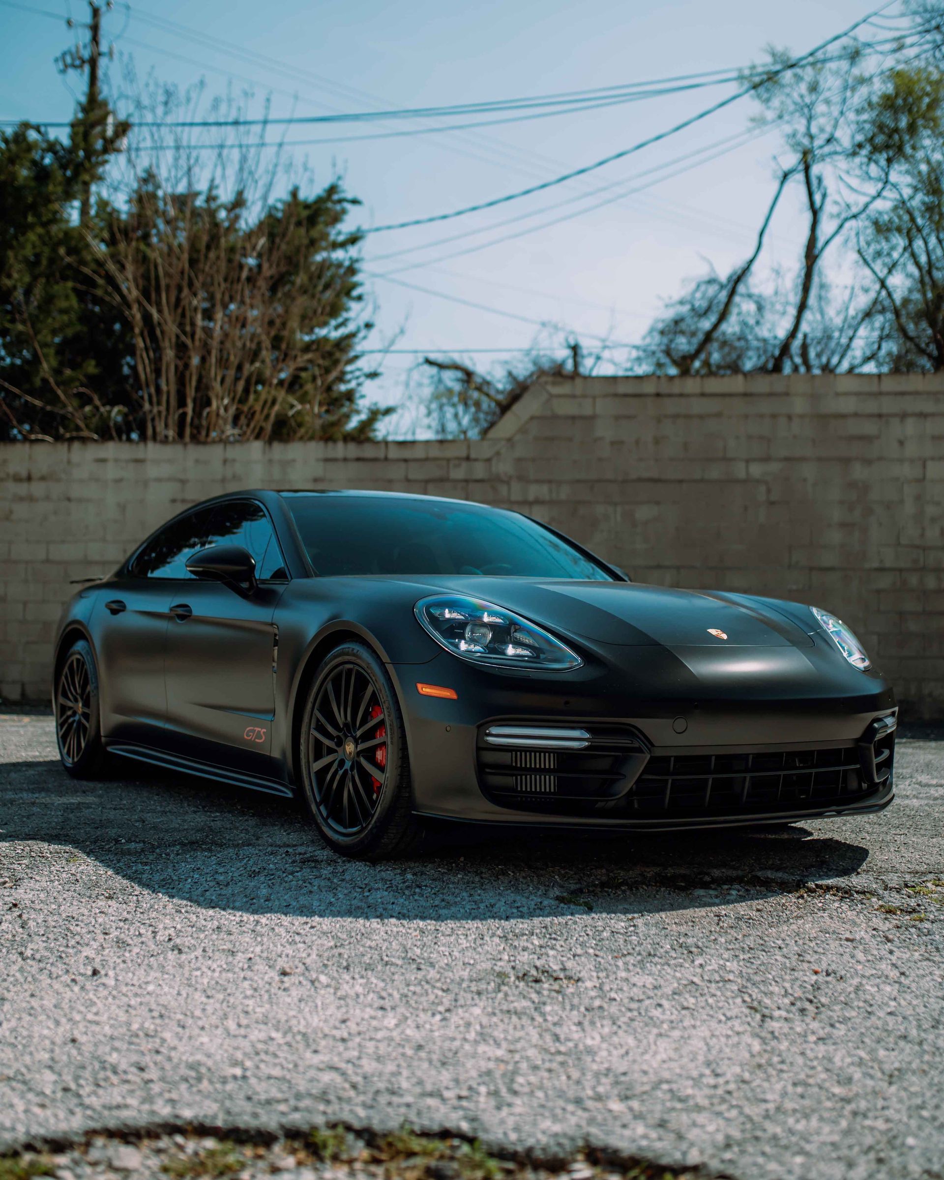 Black sports car parked on gravel in front of a concrete wall, under a blue sky