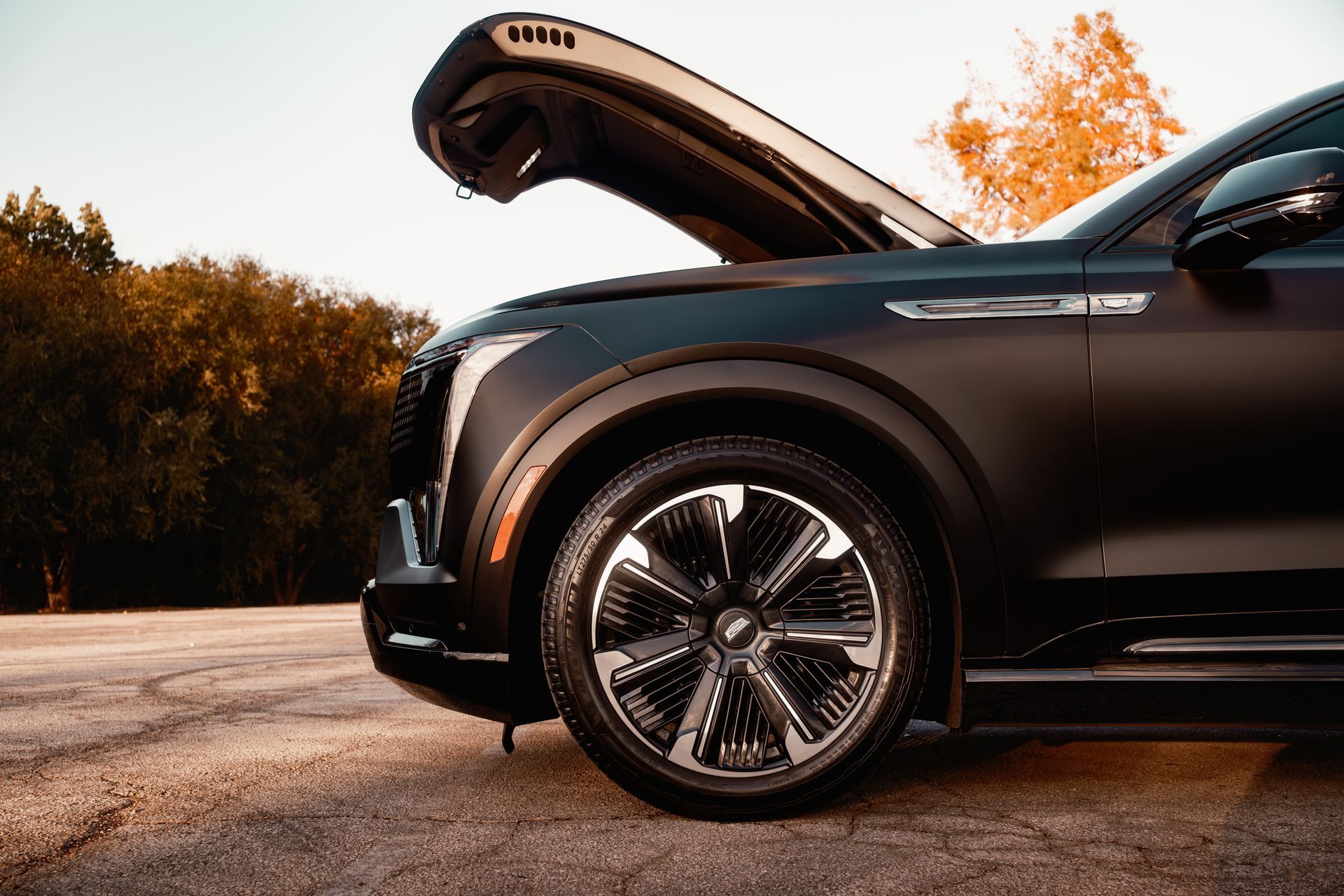 Black SUV with open hood parked outdoors on a road beside autumn trees
