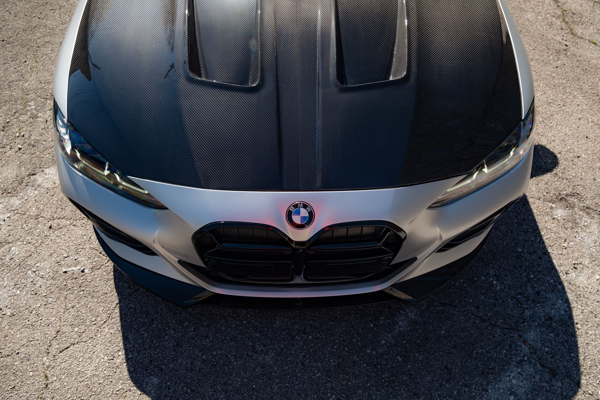 Front view of a silver BMW with a carbon fiber hood parked on sunlit pavement