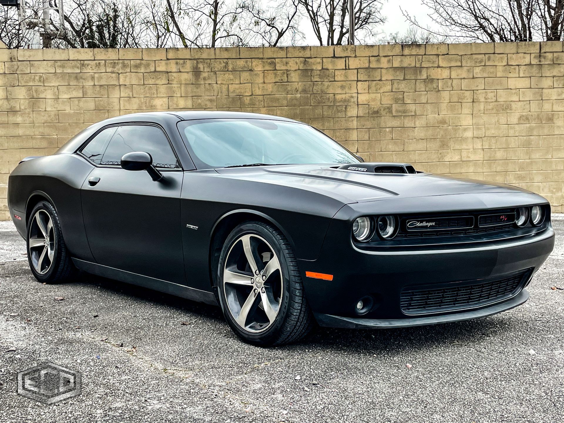 Black Dodge Challenger parked on gravel in front of a tan stone wall