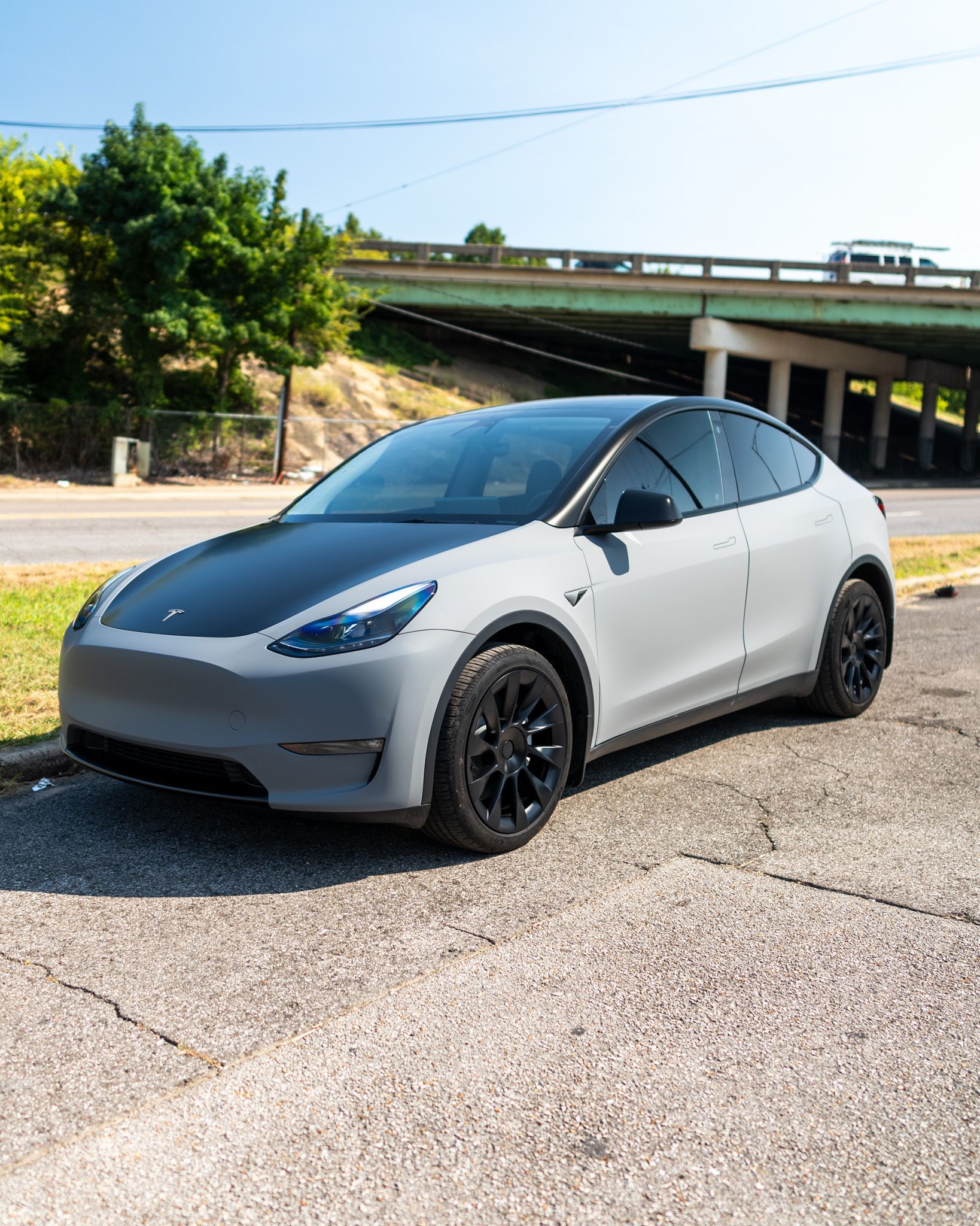 White Tesla parked on a road under an overpass, with black wheels and a sunny sky.
