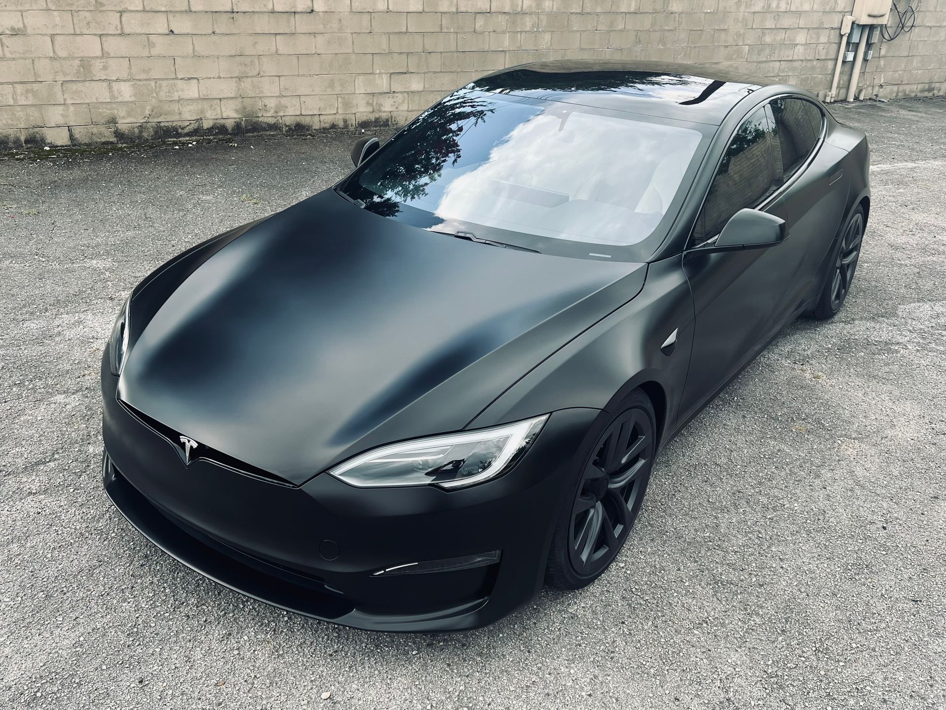 Black sedan with matte finish parked on a gravel lot beside a brick wall.