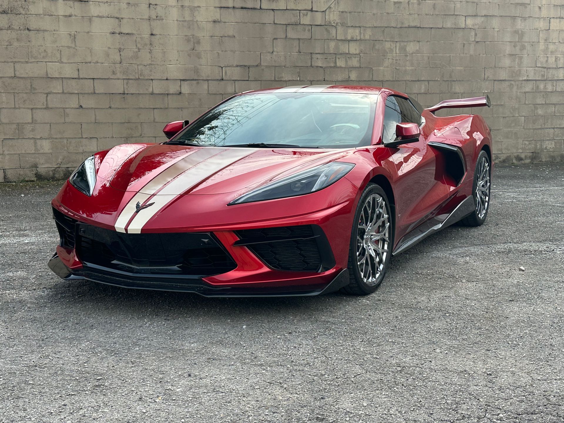 Red Chevrolet Corvette with white racing stripes parked on gravel in front of a concrete wall