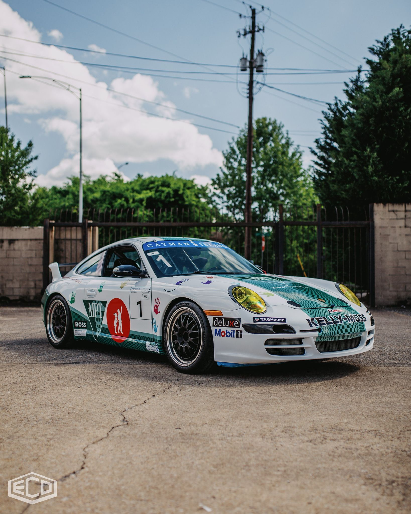 White and green race car with sponsor decals parked on a concrete lot under a partly cloudy sky