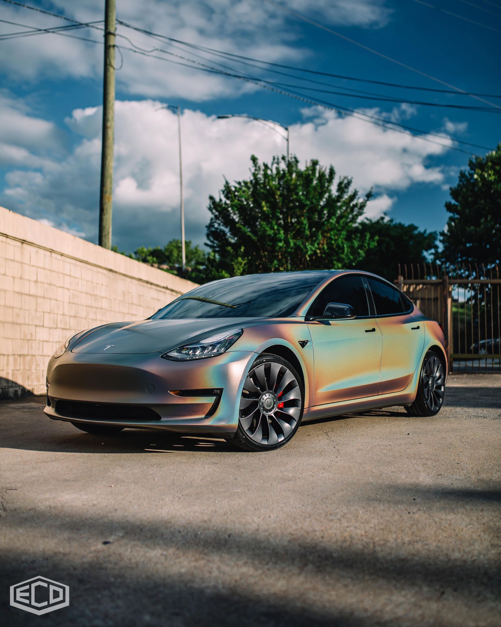 Shiny iridescent sedan parked on a sunny street beside a wall, with trees and blue sky behind it