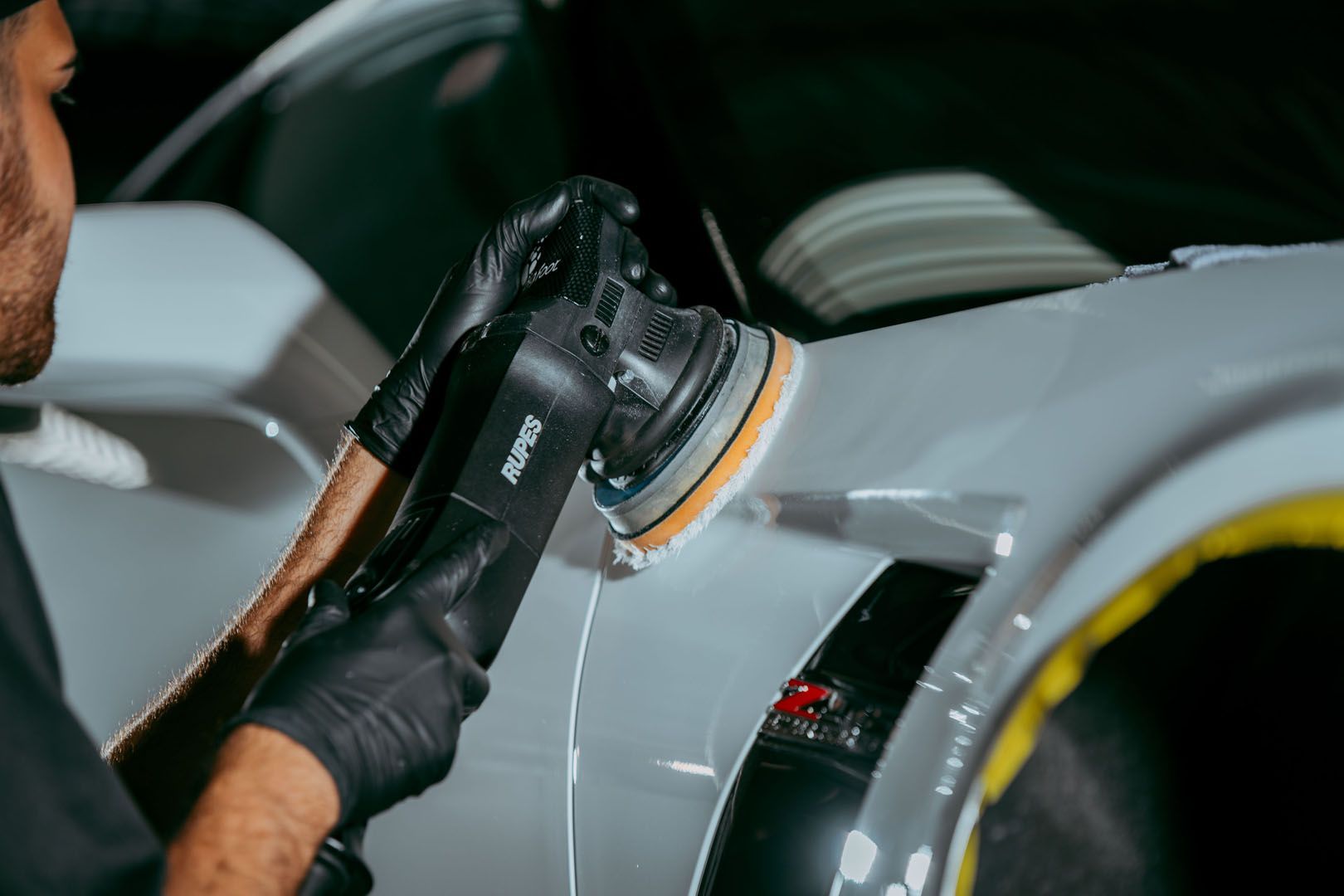 Person polishing a silver car with a buffer, wearing black gloves in a workshop