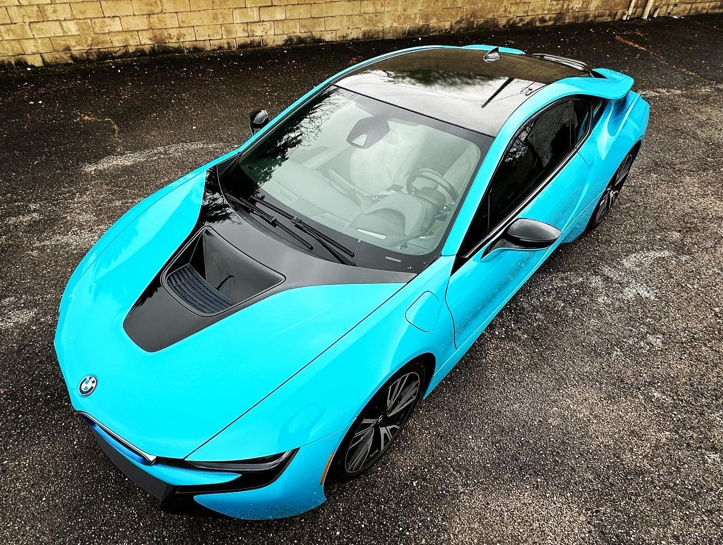 Bright turquoise sports car parked on a wet street beside a stone wall.