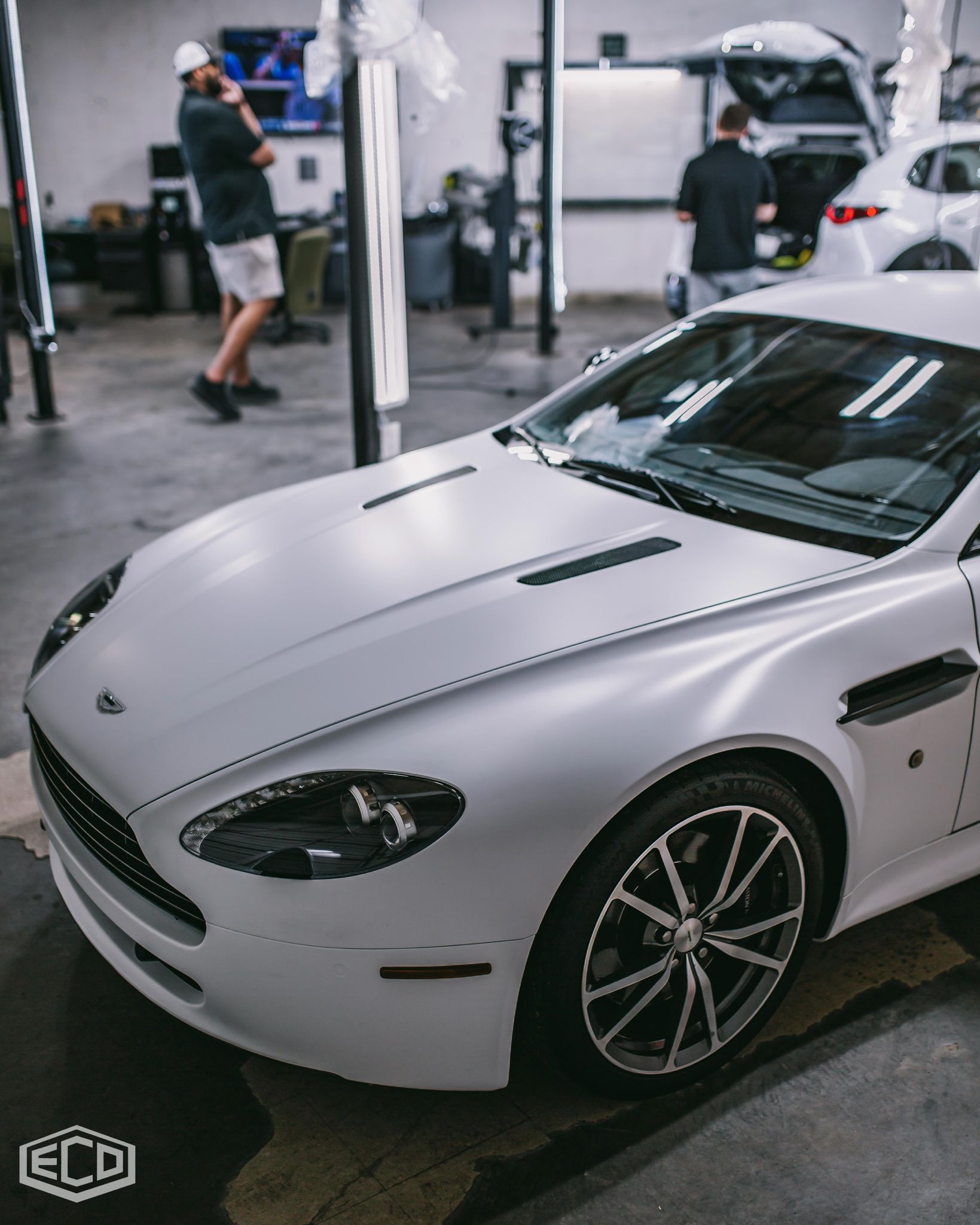 White sports car in an auto workshop, with mechanics and another car in the background