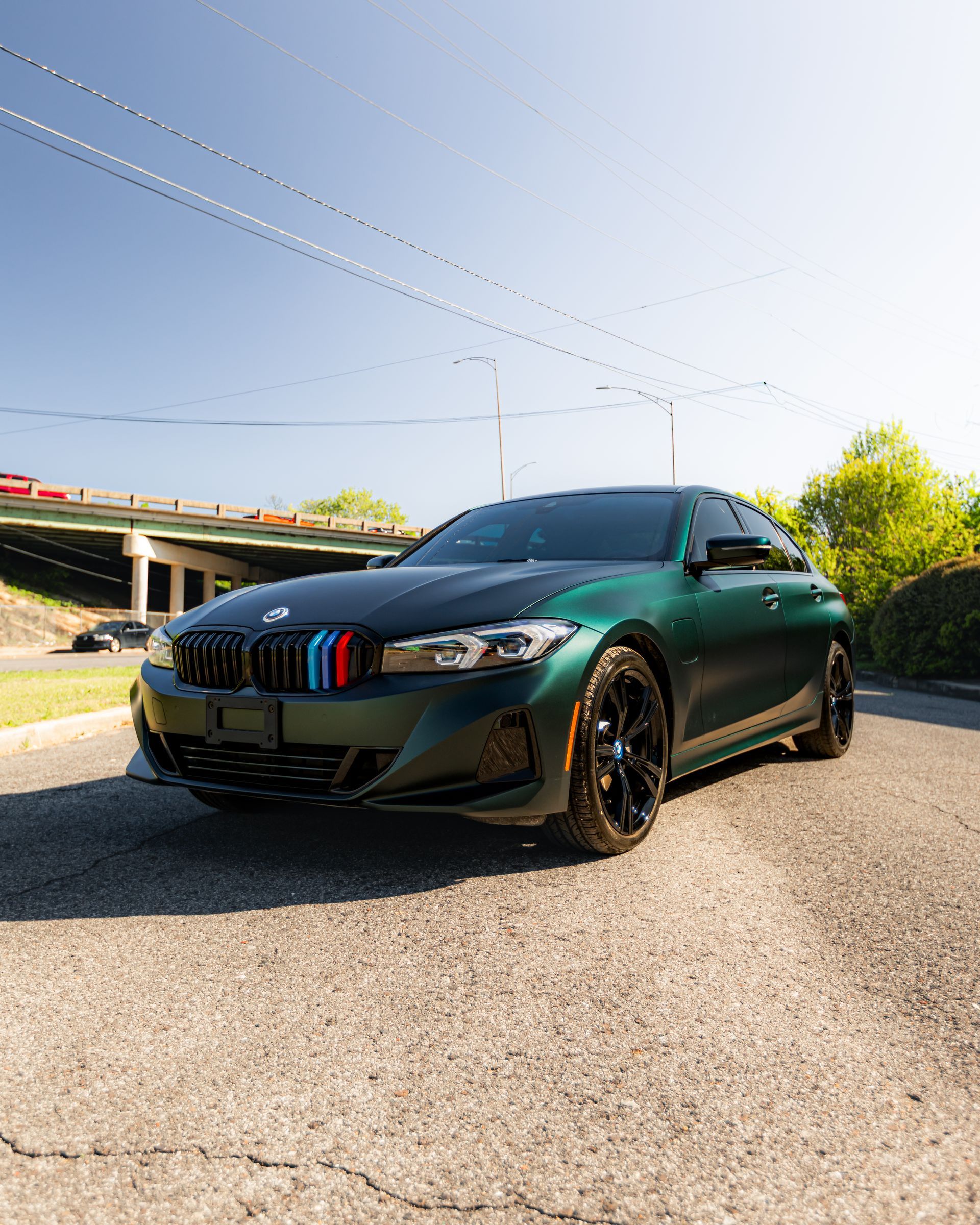 Dark green BMW sedan parked on a sunny street near an overpass, front three-quarter view