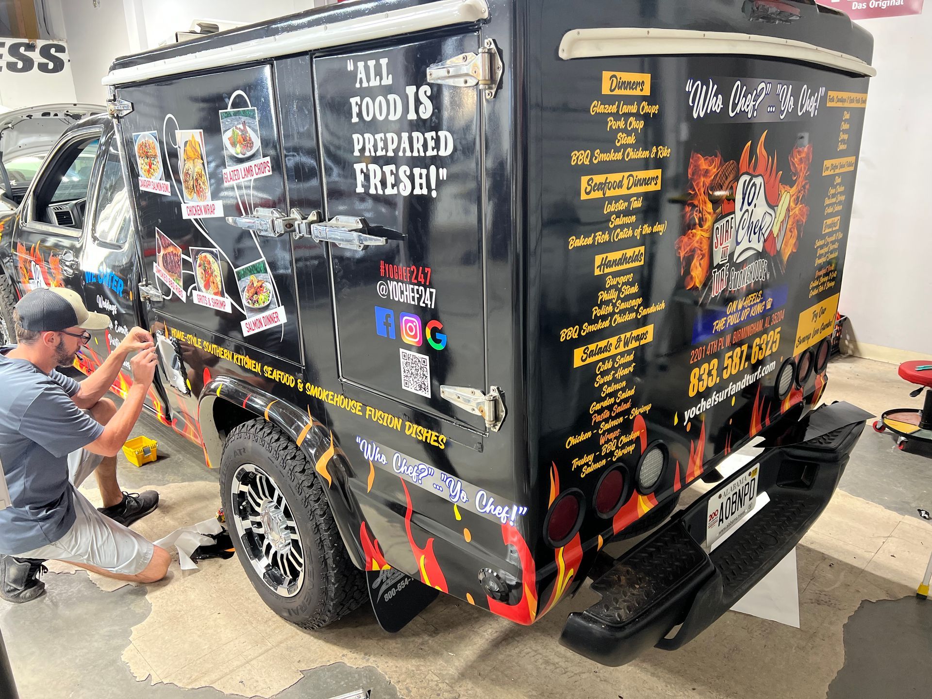 Custom black food truck with colorful menu graphics while a person crouches beside it painting wheels