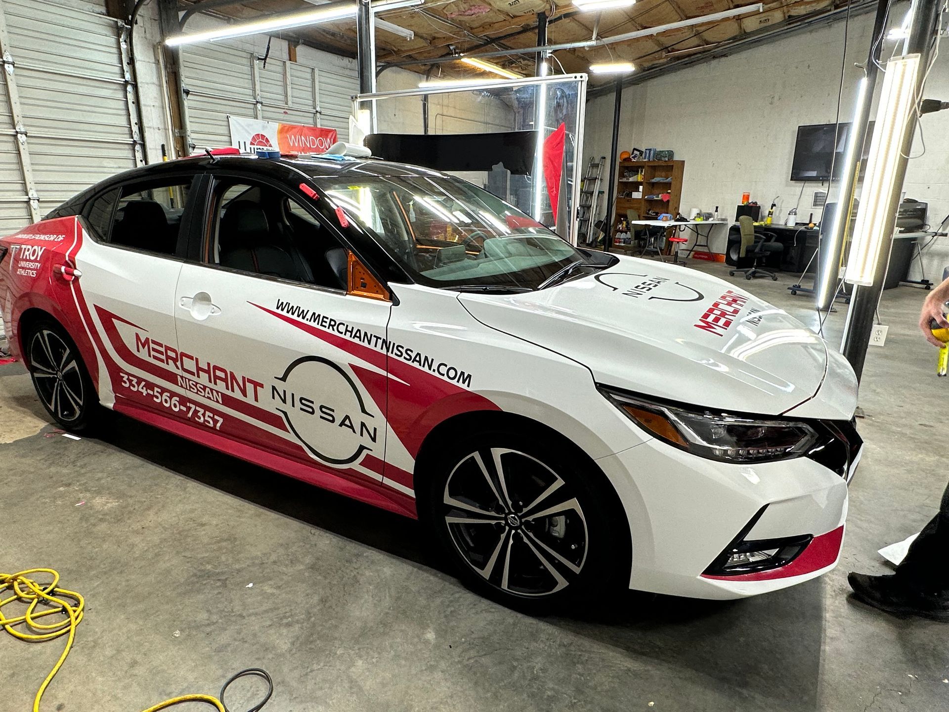 White and red Nissan sedan with branding, parked in an auto shop garage under repair lifts
