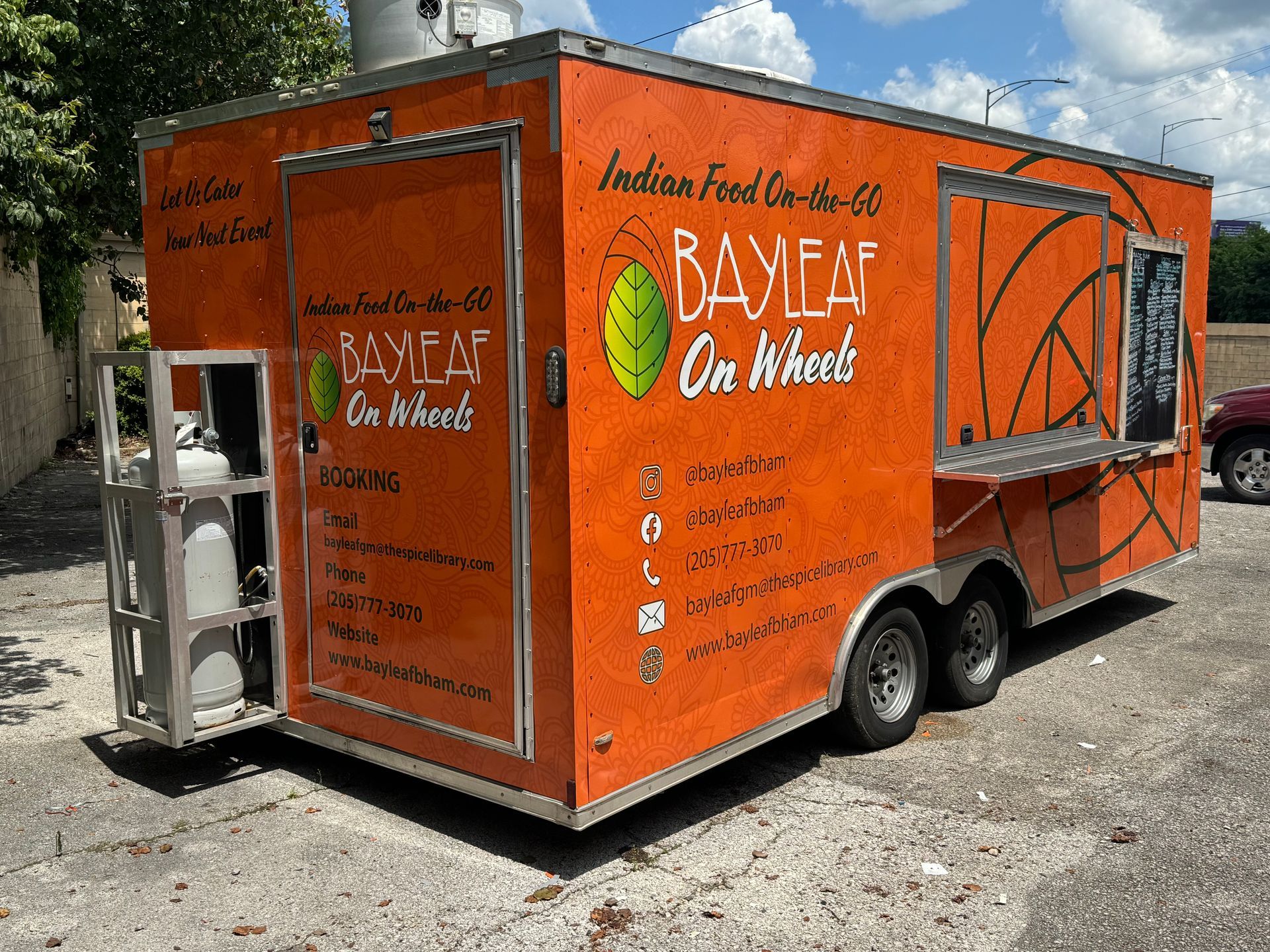 Orange food trailer reading “Bayah On Wheels” parked outdoors on gravel.